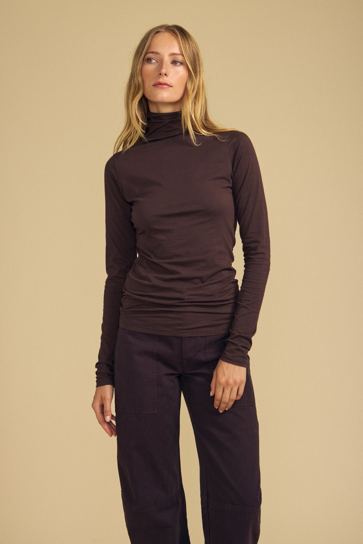 A person with long blonde hair wears the TALISIA TEE by Velvet by Graham & Spencer in dark brown, paired with matching dark brown pants, standing against a beige background.