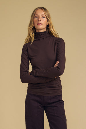 A woman stands with arms crossed against a plain beige background, wearing the TALISIA TEE by Velvet by Graham & Spencer—a long-sleeve brown mock neck—paired with brown pants.