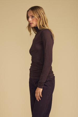 A person stands in profile against a beige background, wearing the TALISIA TEE by Velvet by Graham & Spencer—a fitted brown mock neck turtleneck—paired with dark pants, their arms relaxed at their sides.