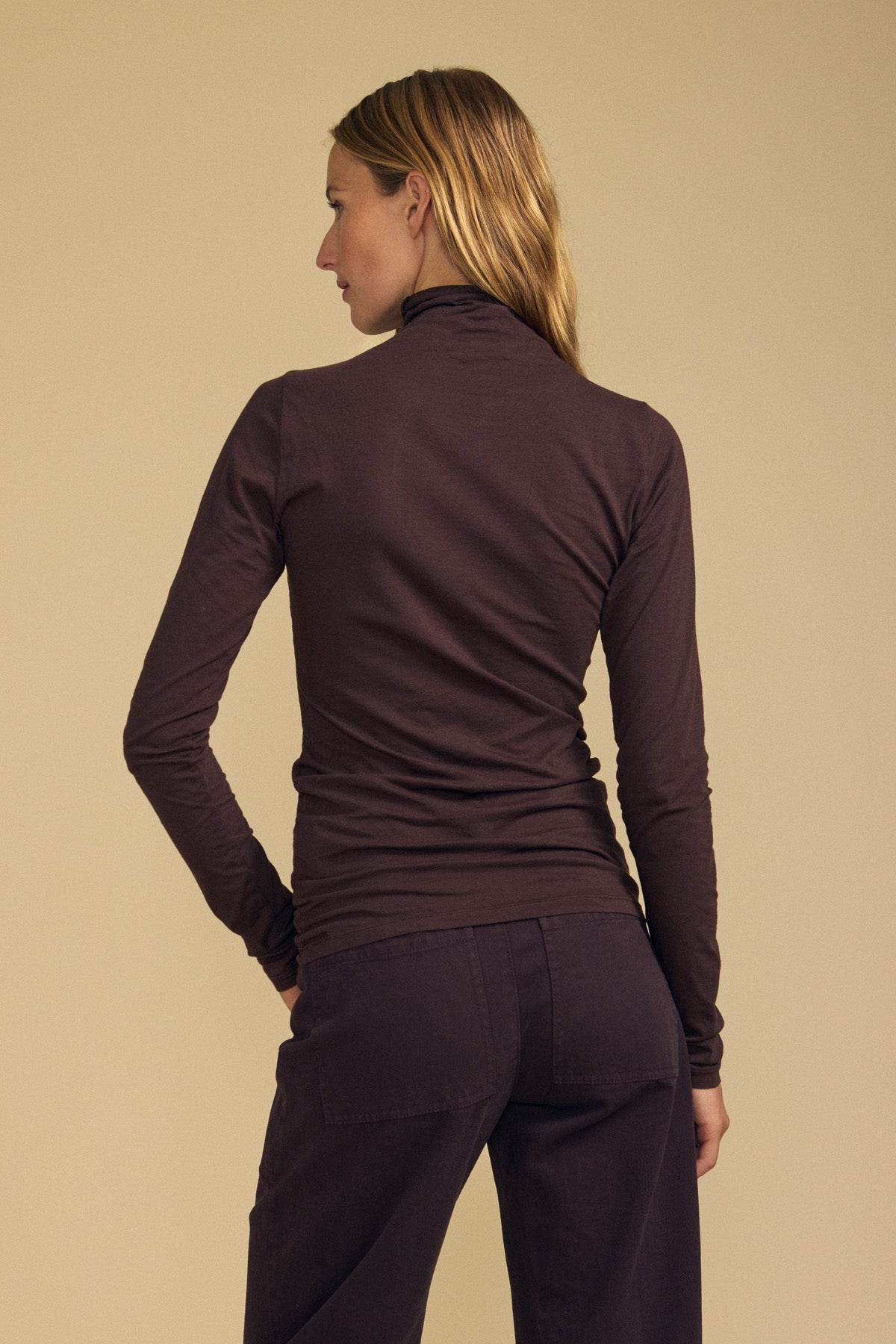 A person with long hair, dressed in the Velvet by Graham & Spencer TALISIA TEE and brown pants, stands facing away from the camera against a beige background.-41964719636673