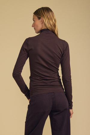 A person with long hair, dressed in the Velvet by Graham & Spencer TALISIA TEE and brown pants, stands facing away from the camera against a beige background.