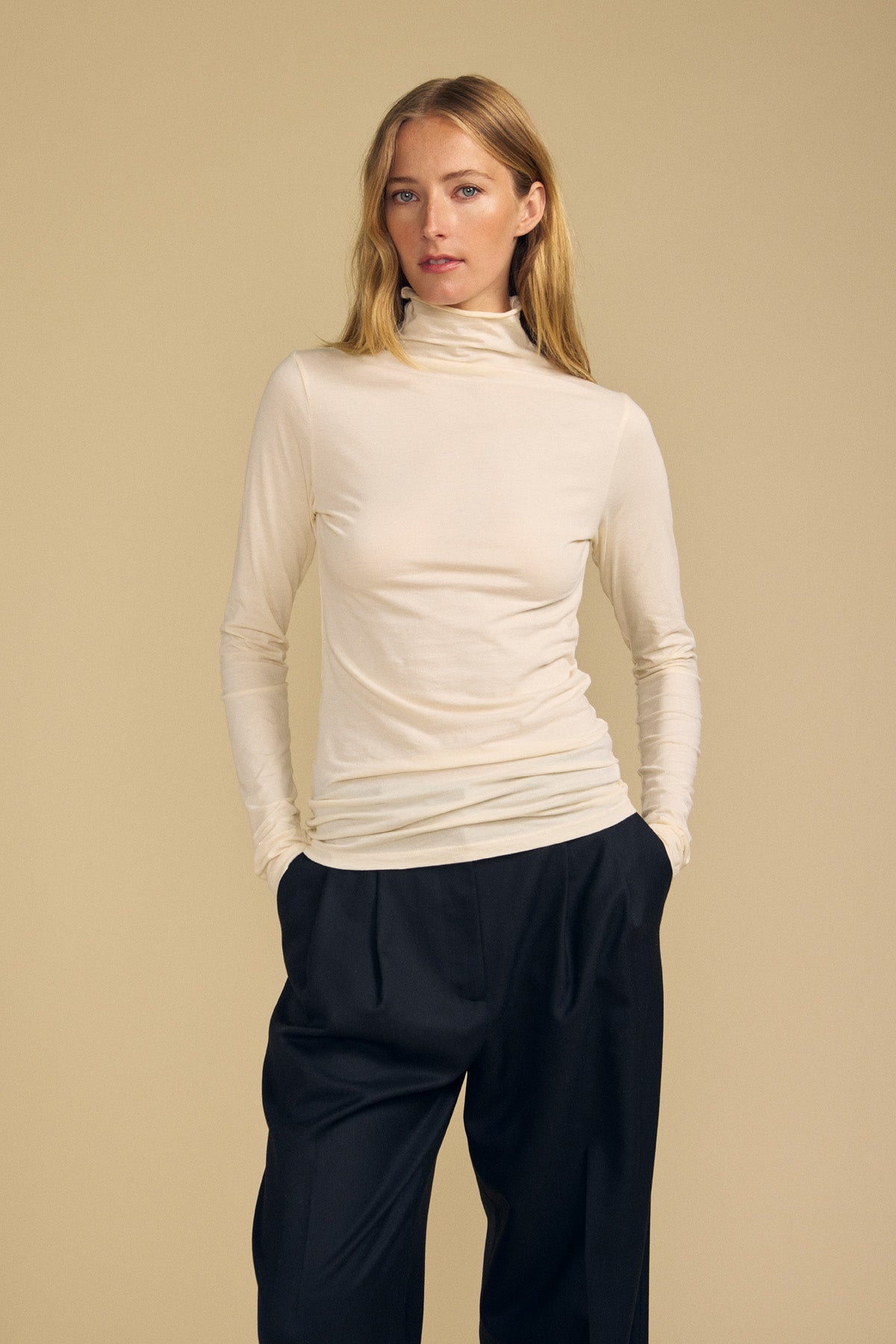 Wearing the TALISIA TEE by Velvet by Graham & Spencer—a cream mock neck long-sleeve top—paired with loose black trousers, she stands with her hands in her pockets against a beige background, embodying a true wardrobe staple.-41964718489793