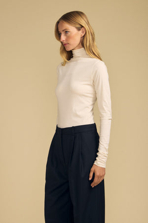 A person stands against a beige background wearing the TALISIA TEE by Velvet by Graham & Spencer—a white mock neck long-sleeve top—paired with high-waisted black trousers, creating a timeless look with this effortless wardrobe staple.