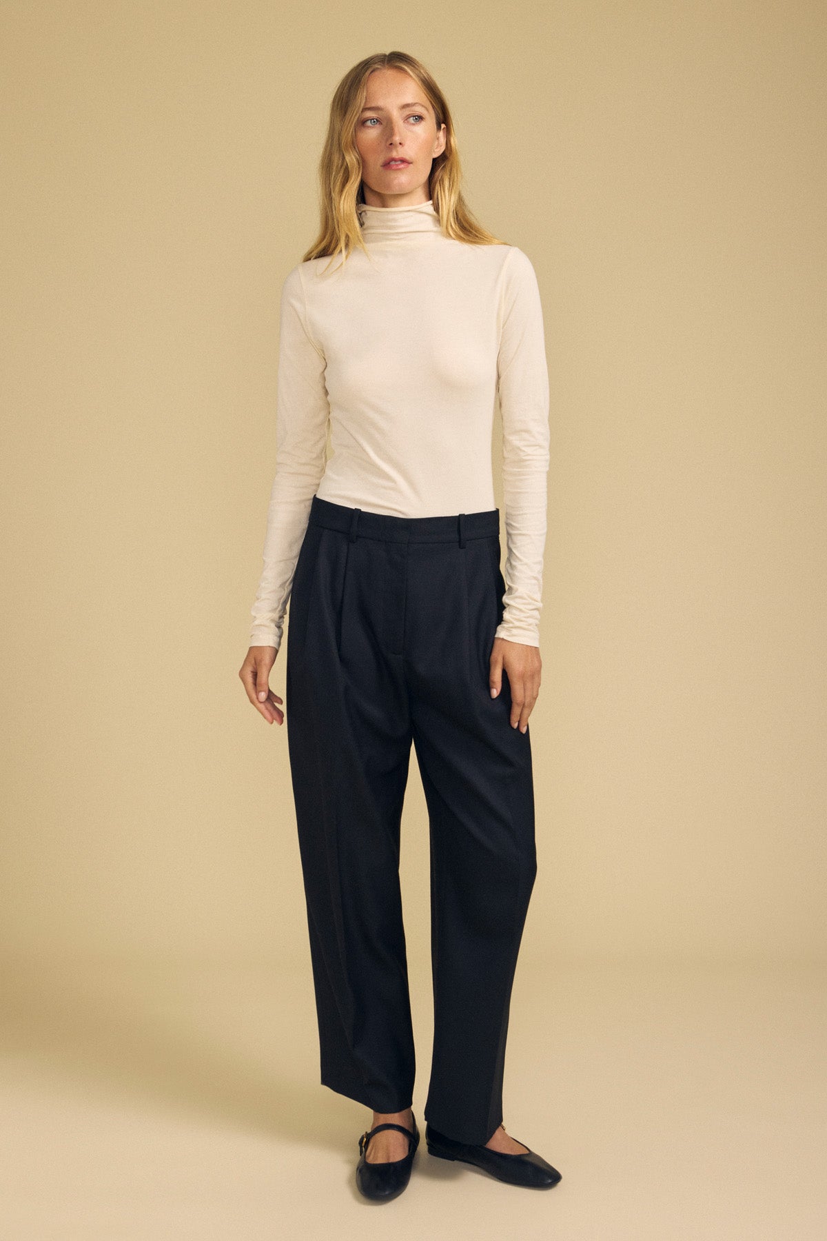 A person stands against a beige background, wearing the TALISIA TEE by Velvet by Graham & Spencer—a cream mock neck turtleneck—paired with black pleated trousers and black flats.
