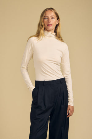 A woman wearing the TALISIA TEE by Velvet by Graham & Spencer—a cream mock neck turtleneck—paired with black pleated trousers stands with one hand in her pocket against a beige background, embodying a true wardrobe staple.