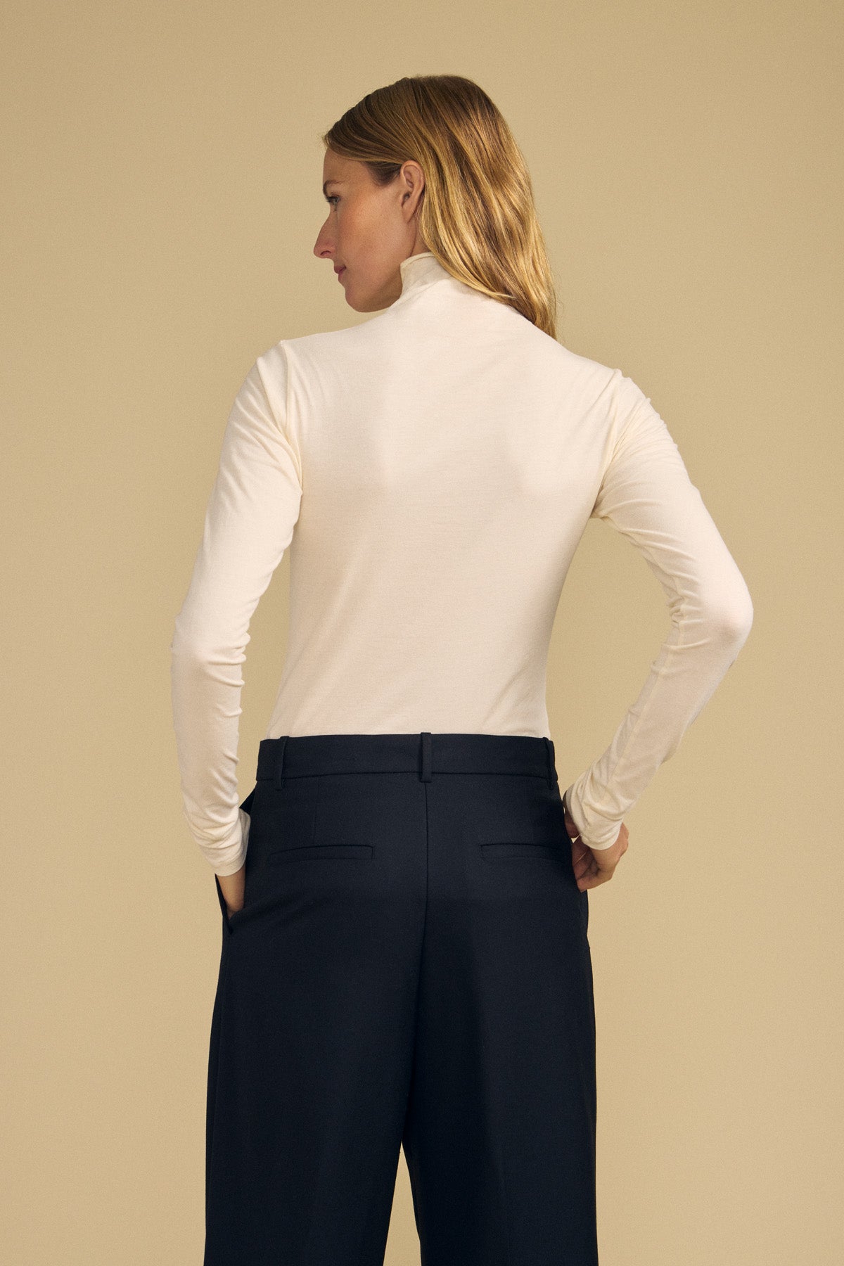 A woman stands with her back to the camera, hands in her pockets, wearing the TALISIA TEE by Velvet by Graham & Spencer—a cream mock neck long-sleeve top—paired with black trousers against a beige background. This effortless ensemble is a true wardrobe staple.