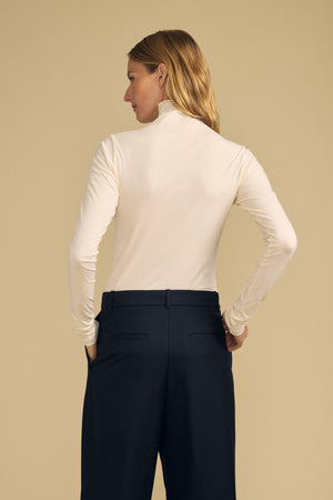 A woman stands with her back to the camera, hands in her pockets, wearing the TALISIA TEE by Velvet by Graham & Spencer—a cream mock neck long-sleeve top—paired with black trousers against a beige background. This effortless ensemble is a true wardrobe staple.