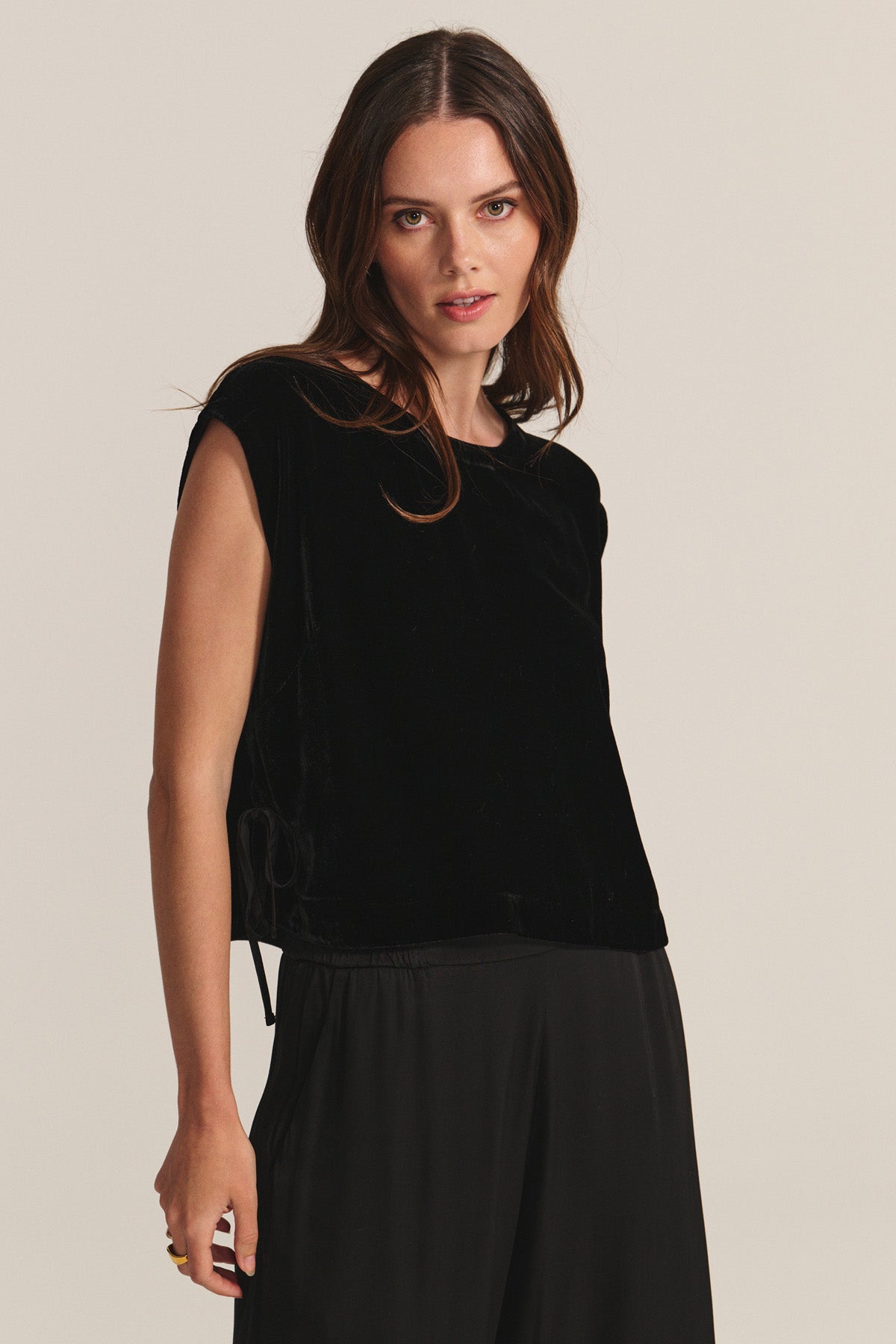 A woman with long brown hair wears the HADLEY TOP by Velvet by Graham & Spencer—a sleeveless, boxy-fit black silk velvet top—paired with loose black pants, standing against a plain light background, ideal for special occasions.