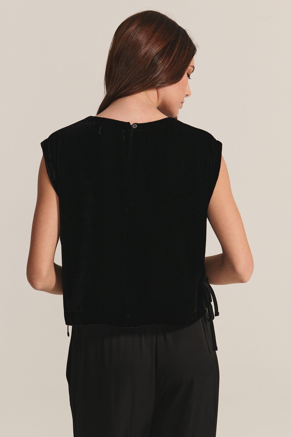 A woman with long brown hair is seen from the back wearing the HADLEY TOP by Velvet by Graham & Spencer—a sleeveless, boxy-fit black silk velvet top—paired with black pants, ideal for special occasions against a plain backdrop.-40263649001665