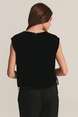 A woman with long brown hair is seen from the back wearing the HADLEY TOP by Velvet by Graham & Spencer—a sleeveless, boxy-fit black silk velvet top—paired with black pants, ideal for special occasions against a plain backdrop.