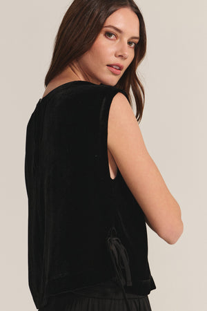 A woman with long brown hair wears the HADLEY TOP—a sleeveless, boxy fit black silk velvet top by Velvet by Graham & Spencer—looking over her shoulder against a neutral background, ideal for special occasions.