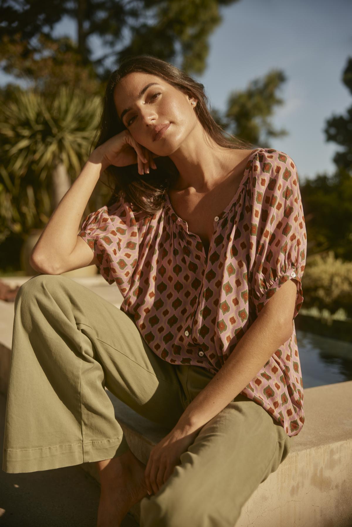   A woman unwinds outside, wearing the HARA TOP from Velvet by Graham & Spencer and green pants, with trees and a pool setting a tranquil scene. 