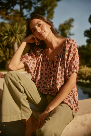 A woman unwinds outside, wearing the HARA TOP from Velvet by Graham & Spencer and green pants, with trees and a pool setting a tranquil scene.