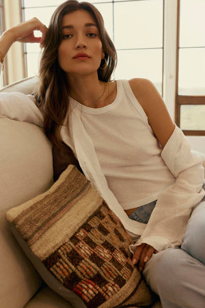 A woman sitting on a couch, resting her head on her hand, dressed in a NATALIA LINEN BUTTON-UP SHIRT and light blue pants, with a thoughtful expression from Velvet by Graham & Spencer.