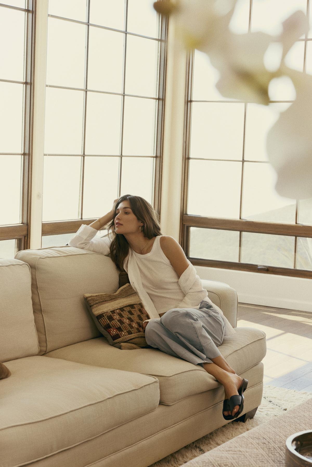   A woman in Velvet by Graham & Spencer's MYA COTTON CANVAS PANT lounges on a beige sofa beside large windows, looking pensive. 