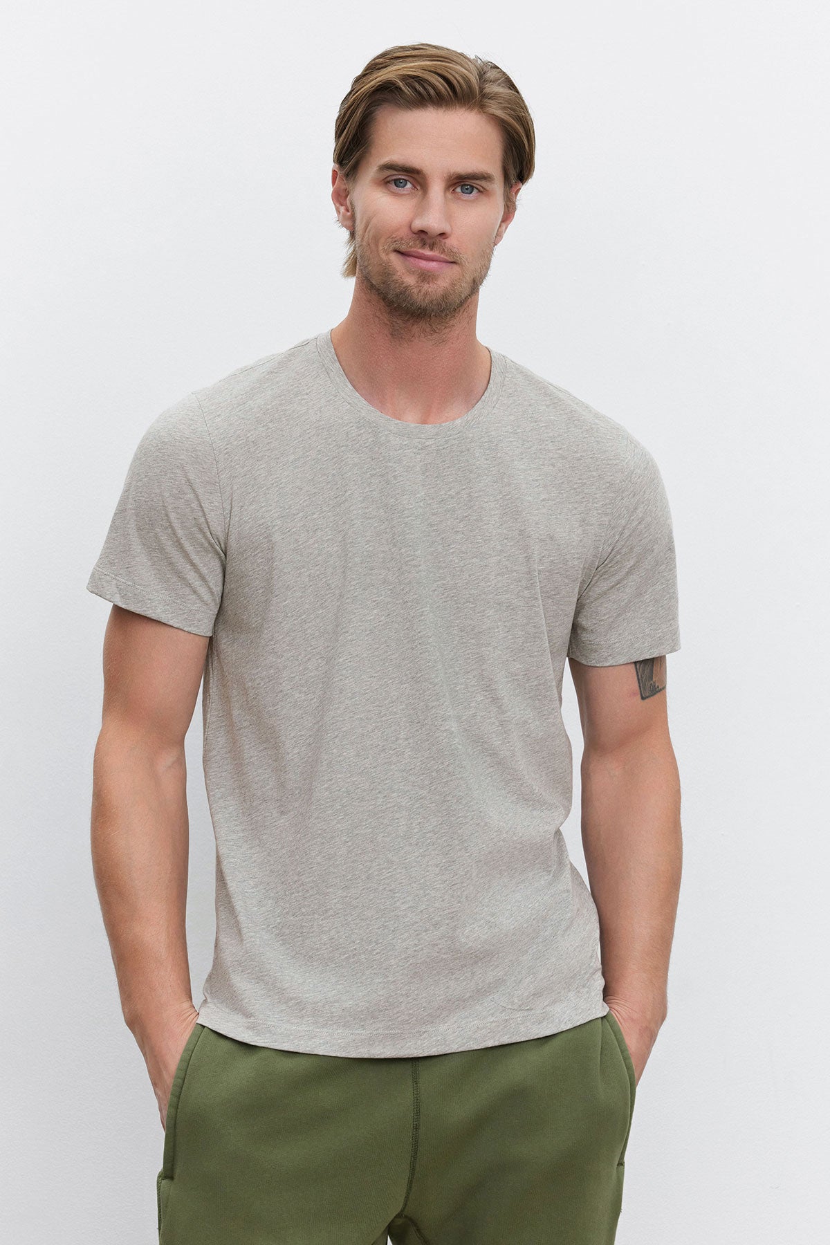 Man with short hair and beard wearing a gray BILLY TEE from Velvet by Graham & Spencer and green pants, standing with hands in pockets against a plain white background.-38537881485505