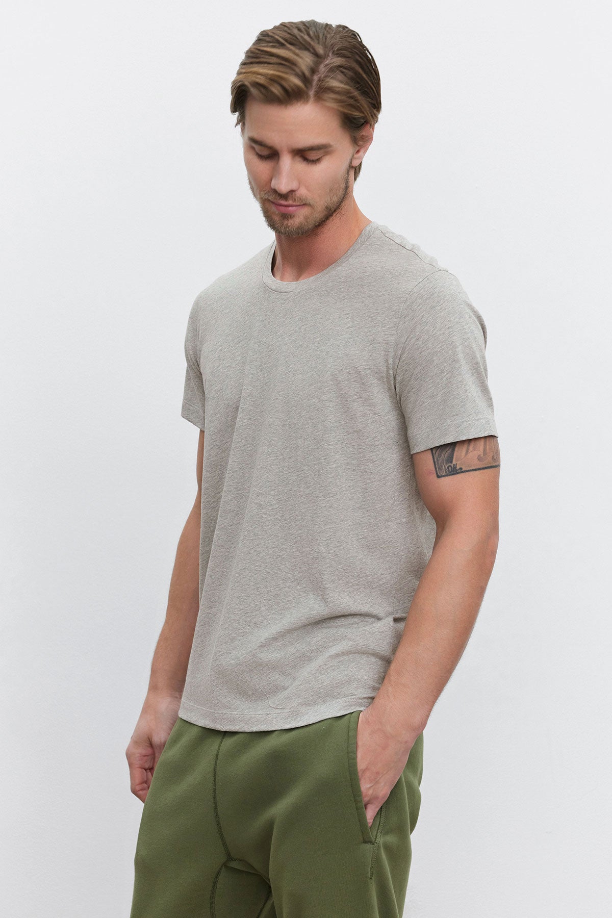 A man in a BILLY TEE by Velvet by Graham & Spencer, featuring a gray heather jersey with a classic crew neckline, pairs it with green pants against a white backdrop, capturing the essence of everyday wear.-38537881518273