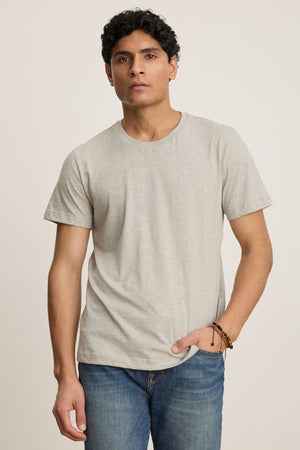 A person with short curly hair models the BILLY TEE by Velvet by Graham & Spencer—a light gray heather jersey crewneck—paired with blue jeans, standing against a plain background for easy everyday style.