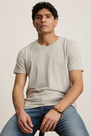 A person with short, curly dark hair wears the BILLY TEE by Velvet by Graham & Spencer in light gray and blue jeans—perfect for everyday wear—while seated on a stool against a plain background.