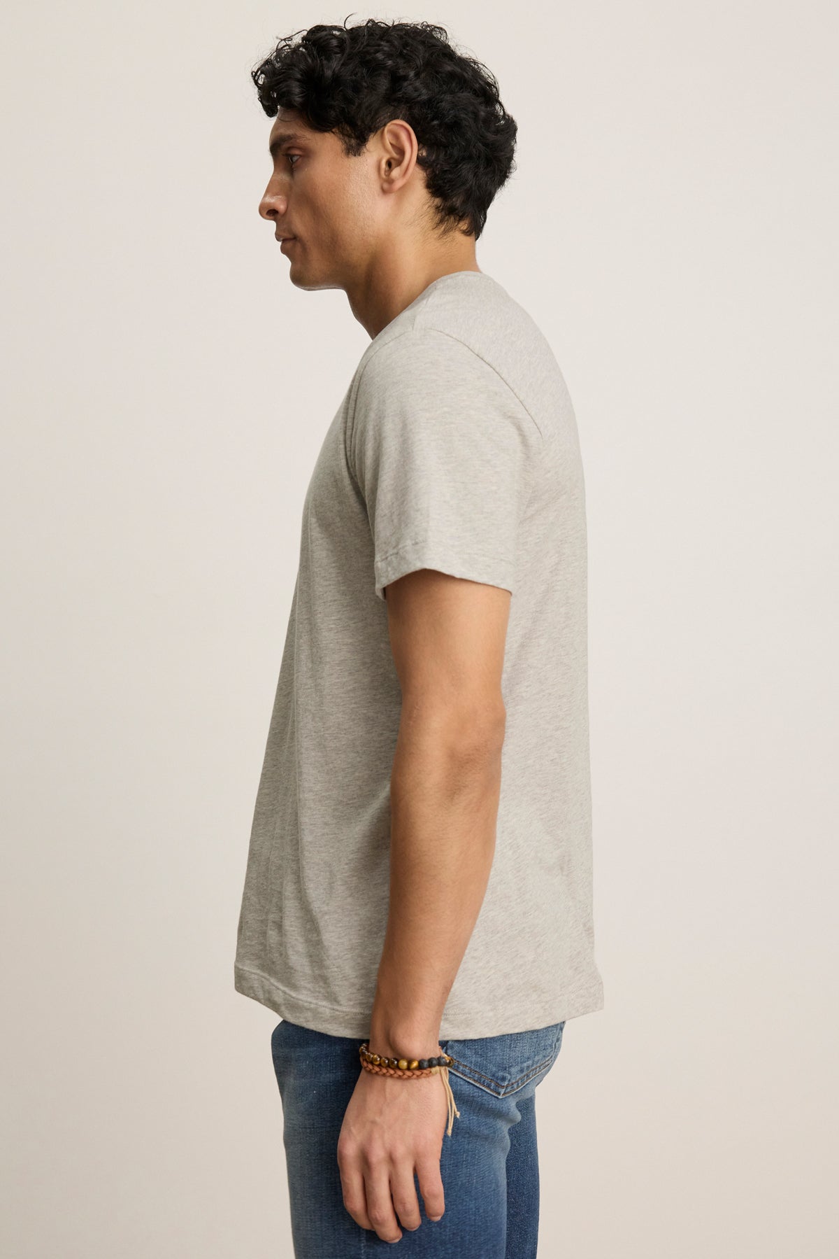 Someone stands in profile against a plain background, wearing Velvet by Graham & Spencer's BILLY TEE—a light gray, soft heather jersey crewneck short-sleeve t-shirt—paired with blue jeans and a beaded bracelet for everyday style.-39543503814849