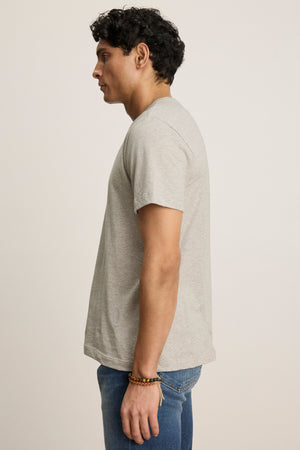 Someone stands in profile against a plain background, wearing Velvet by Graham & Spencer's BILLY TEE—a light gray, soft heather jersey crewneck short-sleeve t-shirt—paired with blue jeans and a beaded bracelet for everyday style.