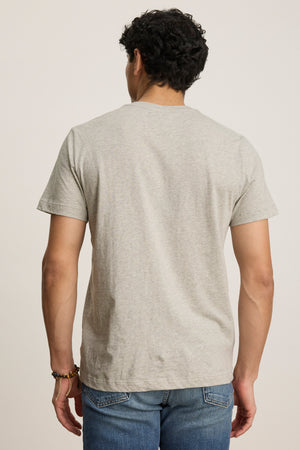 A man with dark curly hair is seen from behind wearing the BILLY TEE by Velvet by Graham & Spencer—a light gray heather jersey short-sleeve crewneck paired with blue jeans, ideal for everyday style against a plain background.
