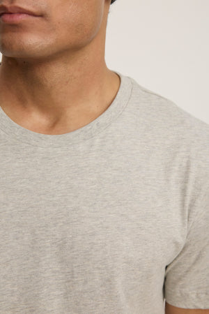 Close-up of a person wearing the BILLY TEE by Velvet by Graham & Spencer in light grey heather, showcasing the crew neck and upper torso—an ideal choice for everyday wear against a plain background.