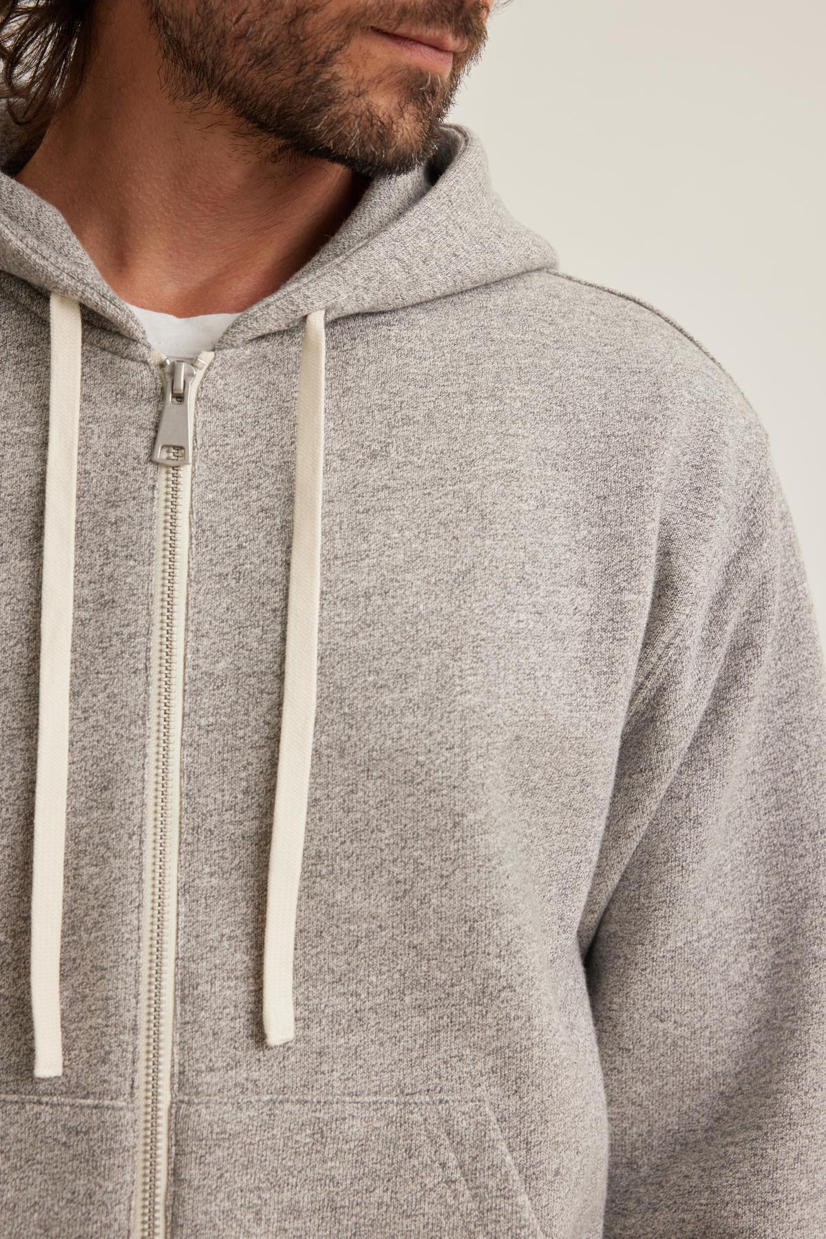   A man wears the DRACO HOODIE by Velvet by Graham & Spencer, a gray zip-up hoodie featuring white drawstrings, with his partially visible head and beard set against a light background. 