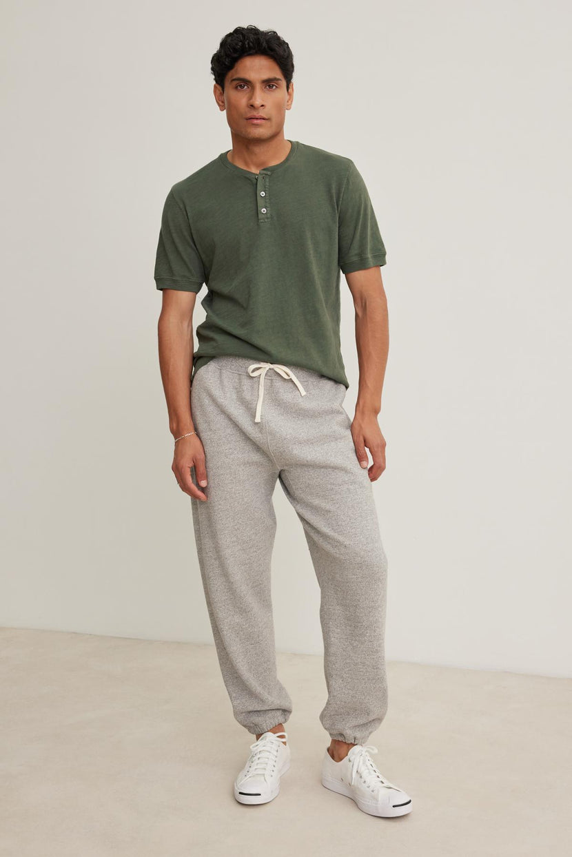 A man stands wearing a green henley shirt, Velvet by Graham & Spencer KEELAN JOGGER in gray, and white sneakers against a plain background.