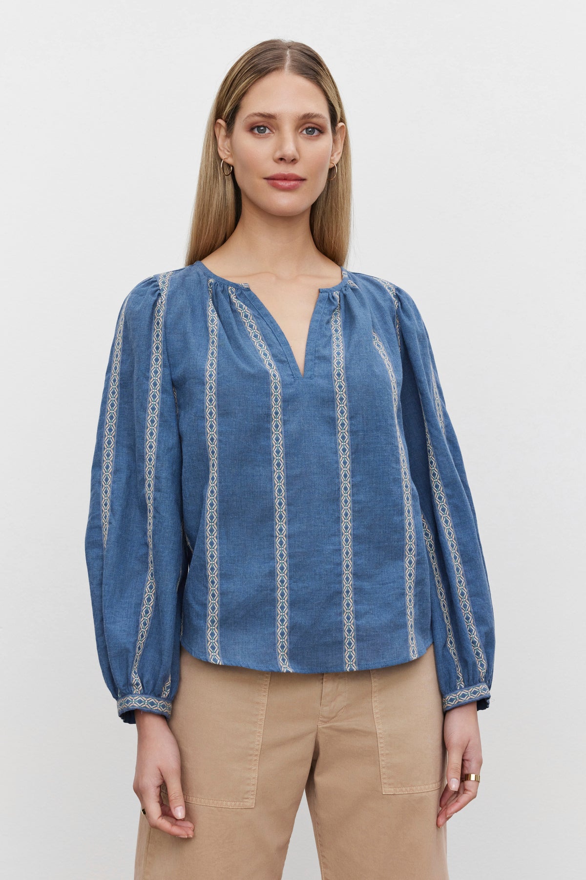   A woman with straight blond hair is wearing a relaxed fit RAYLEE TOP by Velvet by Graham & Spencer in blue, adorned with intricate white patterns, and beige pants. She is standing against a plain white background. 