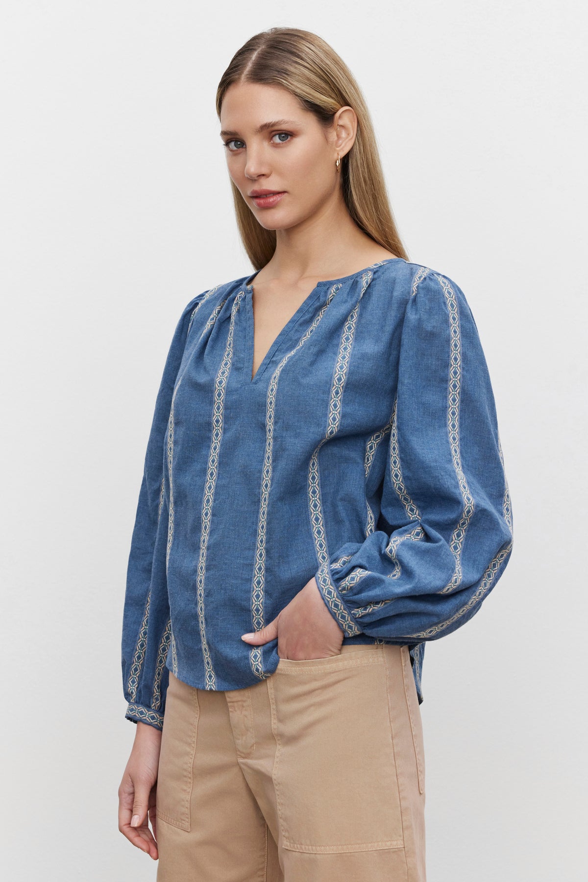   A person with long hair wearing a Velvet by Graham & Spencer RAYLEE TOP in blue with patterned stripes and beige pants stands against a plain white background, the relaxed fit complemented by a trendy split neck. 