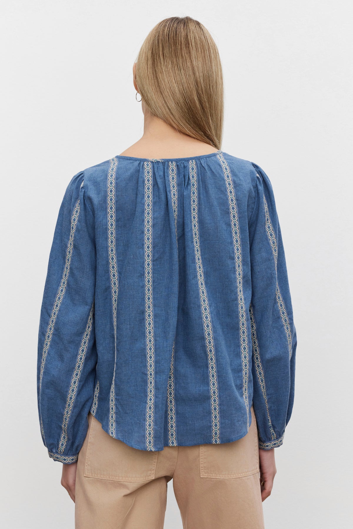   A person with long blonde hair is seen from the back, wearing the RAYLEE TOP from Velvet by Graham & Spencer, which showcases a blue blouse with white patterned vertical stripes and beige pants. The RAYLEE TOP features a relaxed fit and a split neck, giving it an effortlessly stylish look. 