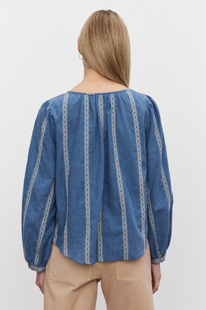 A person with long blonde hair is seen from the back, wearing the RAYLEE TOP from Velvet by Graham & Spencer, which showcases a blue blouse with white patterned vertical stripes and beige pants. The RAYLEE TOP features a relaxed fit and a split neck, giving it an effortlessly stylish look.