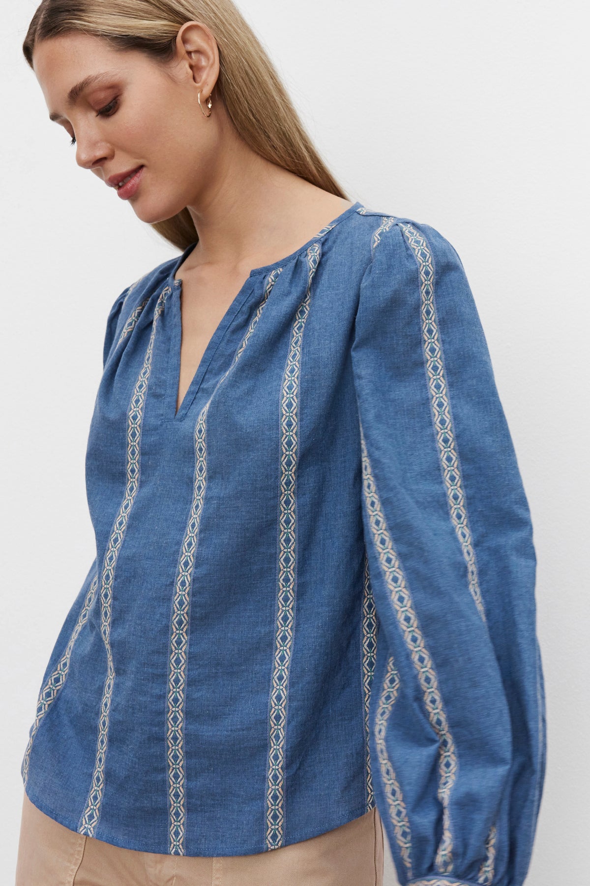   A person with long hair is wearing the Velvet by Graham & Spencer RAYLEE TOP, which features blue heathered striped jacquard cotton and white geometric embroidery. They are looking downward with a neutral expression against a plain white background. 