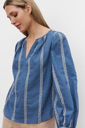 A person with long hair is wearing the Velvet by Graham & Spencer RAYLEE TOP, which features blue heathered striped jacquard cotton and white geometric embroidery. They are looking downward with a neutral expression against a plain white background.