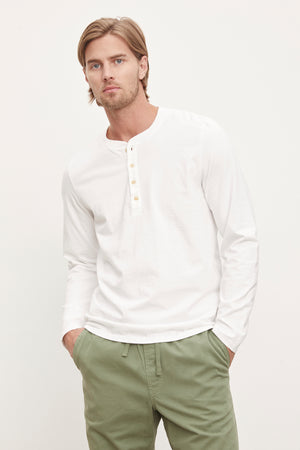 Man with blond hair wearing a white long-sleeve Velvet by Graham & Spencer HOLT HENLEY henley shirt with a four-button placket and green pants, standing against a plain background.