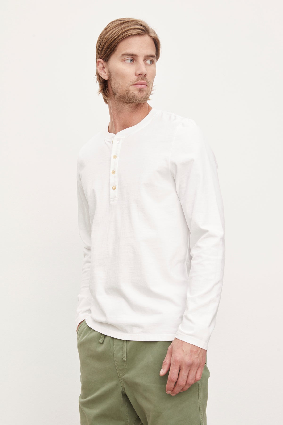   A man in a white long-sleeve cotton velvet by Graham & Spencer HOLT HENLEY shirt with a four-button placket and green pants, looking to the side against a plain background. 