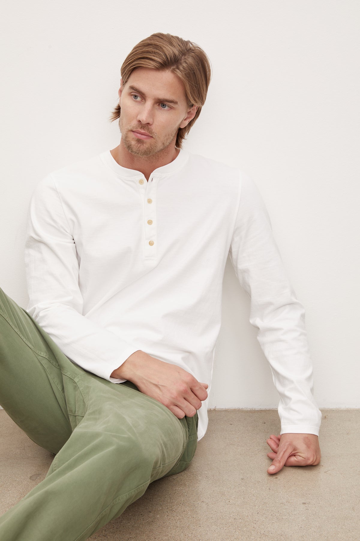   A young man with blond hair wearing a white long-sleeve cotton Velvet by Graham & Spencer HOLT HENLEY henley shirt and olive green pants, sitting against a white wall, looking to his left. 