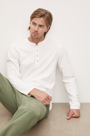 A young man with blond hair wearing a white long-sleeve cotton Velvet by Graham & Spencer HOLT HENLEY henley shirt and olive green pants, sitting against a white wall, looking to his left.