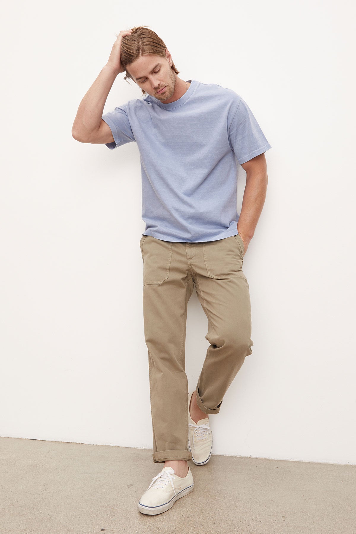   A man in a Velvet by Graham & Spencer Jacobi Heavy Jersey Crew Neck Tee and khaki trousers leans against a white wall, appearing contemplative or relaxed, with one hand resting on his head. 