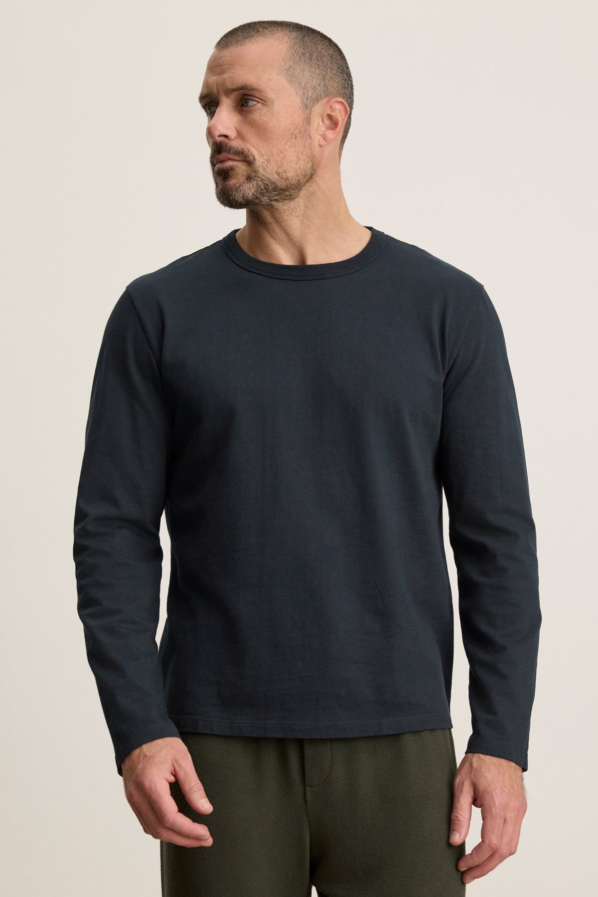 A man with short hair and a beard wears the Velvet by Graham & Spencer BRADY TEE in midweight jersey with dark pants, standing against a neutral background—an ideal choice for everyday style.