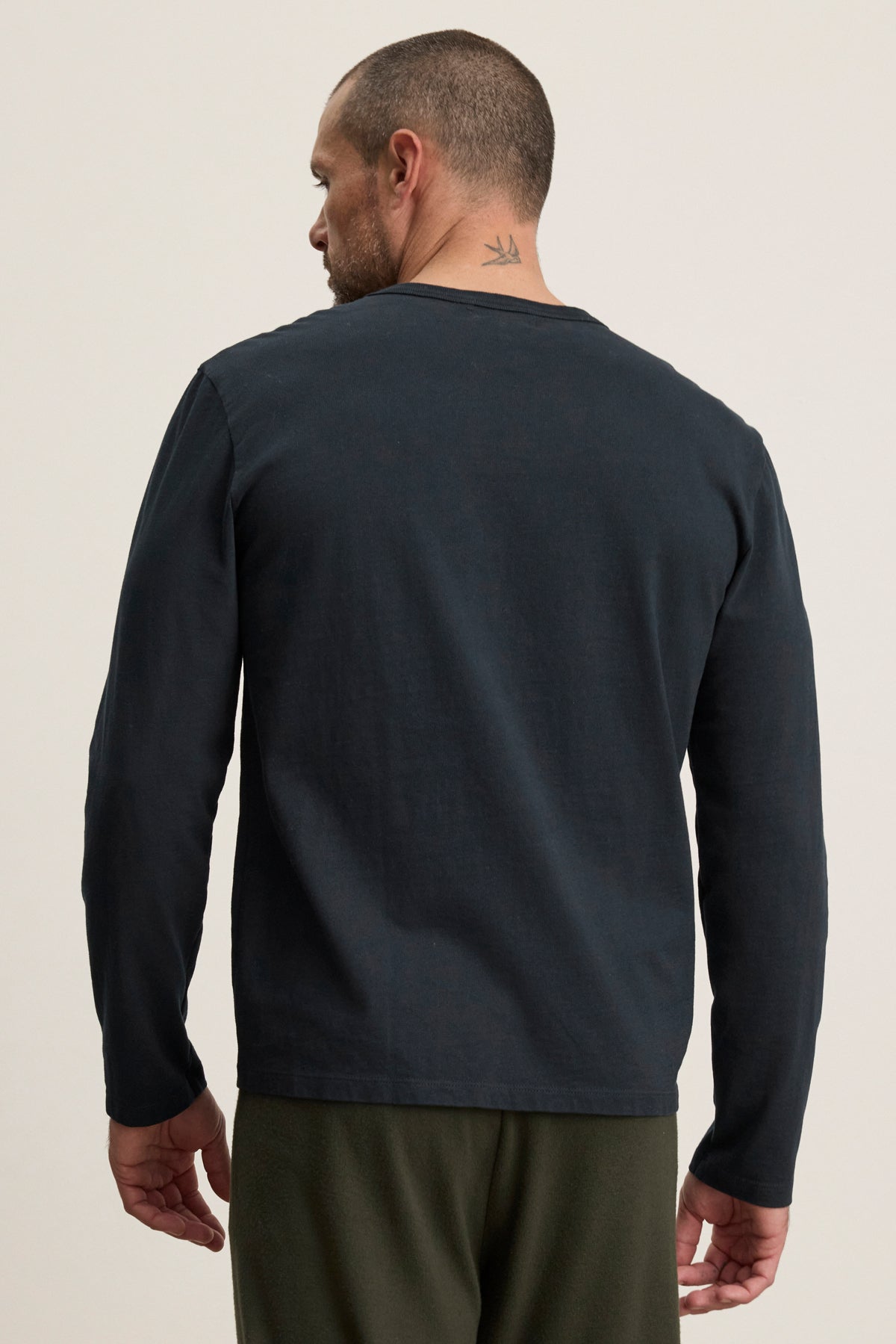 A man with short hair and a bird tattoo on his neck is seen from the back wearing the BRADY TEE by Velvet by Graham & Spencer— a dark, tailored fit, midweight jersey tee, styled with green pants for effortless everyday wear.-40087430136001