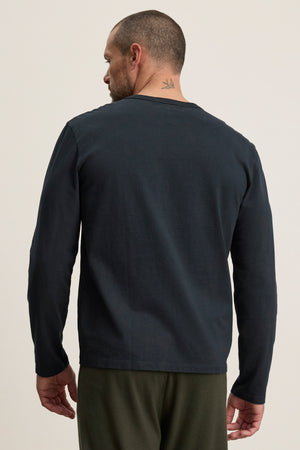 A man with short hair and a bird tattoo on his neck is seen from the back wearing the BRADY TEE by Velvet by Graham & Spencer— a dark, tailored fit, midweight jersey tee, styled with green pants for effortless everyday wear.