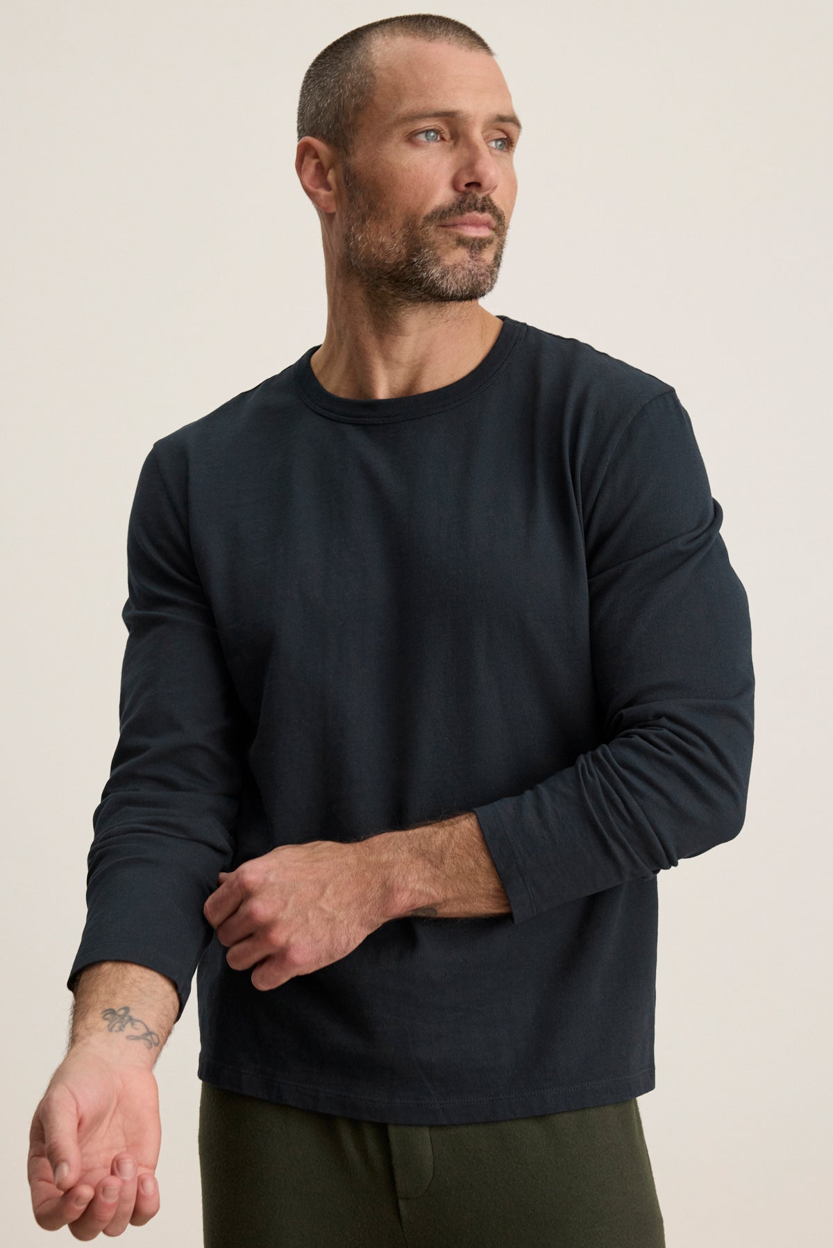 A man with short hair and a beard, wearing the Velvet by Graham & Spencer BRADY TEE and dark pants, looks to the side while adjusting his sleeve—ideal for effortless everyday wear.-40087430070465