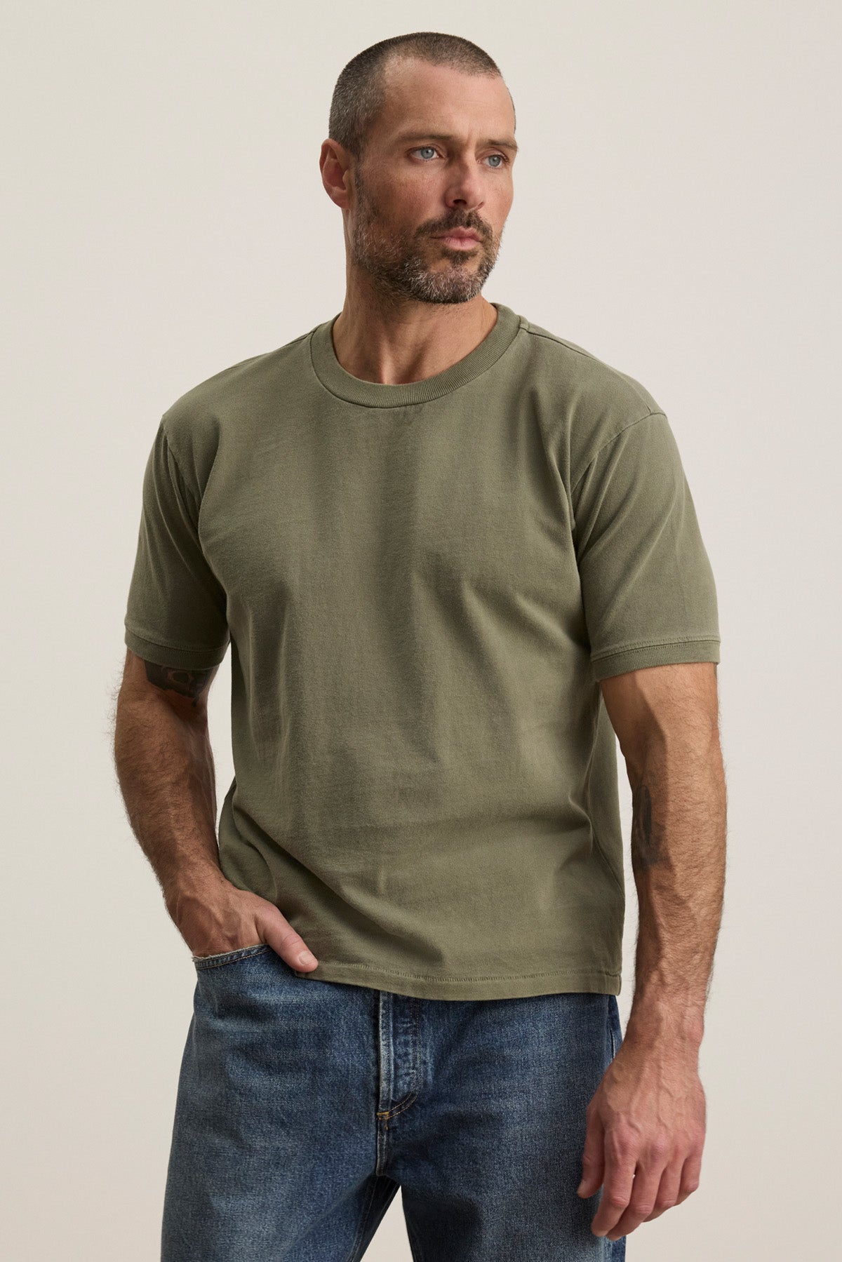 A man with short hair and a beard wears the ELIAS TEE by Velvet by Graham & Spencer in olive green with a classic crew neckline and blue jeans, standing against a plain light background—a timeless wardrobe essential.-40452479516865
