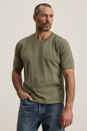 A man with short hair and a beard wears the ELIAS TEE by Velvet by Graham & Spencer in olive green with a classic crew neckline and blue jeans, standing against a plain light background—a timeless wardrobe essential.