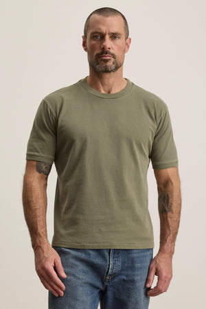 A man with short hair and a beard wears the ELIAS TEE by Velvet by Graham & Spencer—an olive green heavy jersey crew neck—paired with blue jeans, standing against a plain light background.