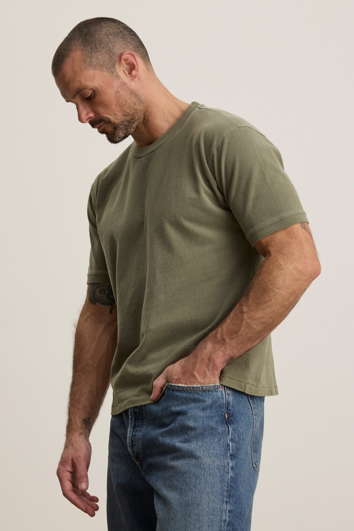 A man with short hair and a beard wears the ELIAS TEE by Velvet by Graham & Spencer, an olive green heavy jersey crewneck t-shirt, paired with blue jeans—creating a classic everyday look against a plain background.