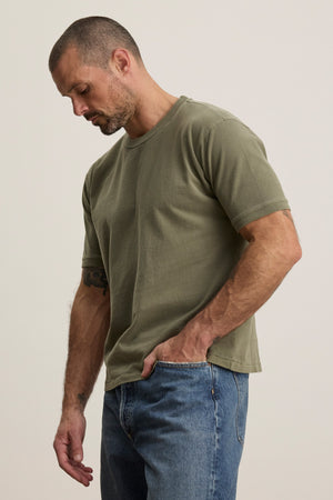 A man with short hair and a beard wears the ELIAS TEE by Velvet by Graham & Spencer, an olive green heavy jersey crewneck t-shirt, paired with blue jeans—creating a classic everyday look against a plain background.
