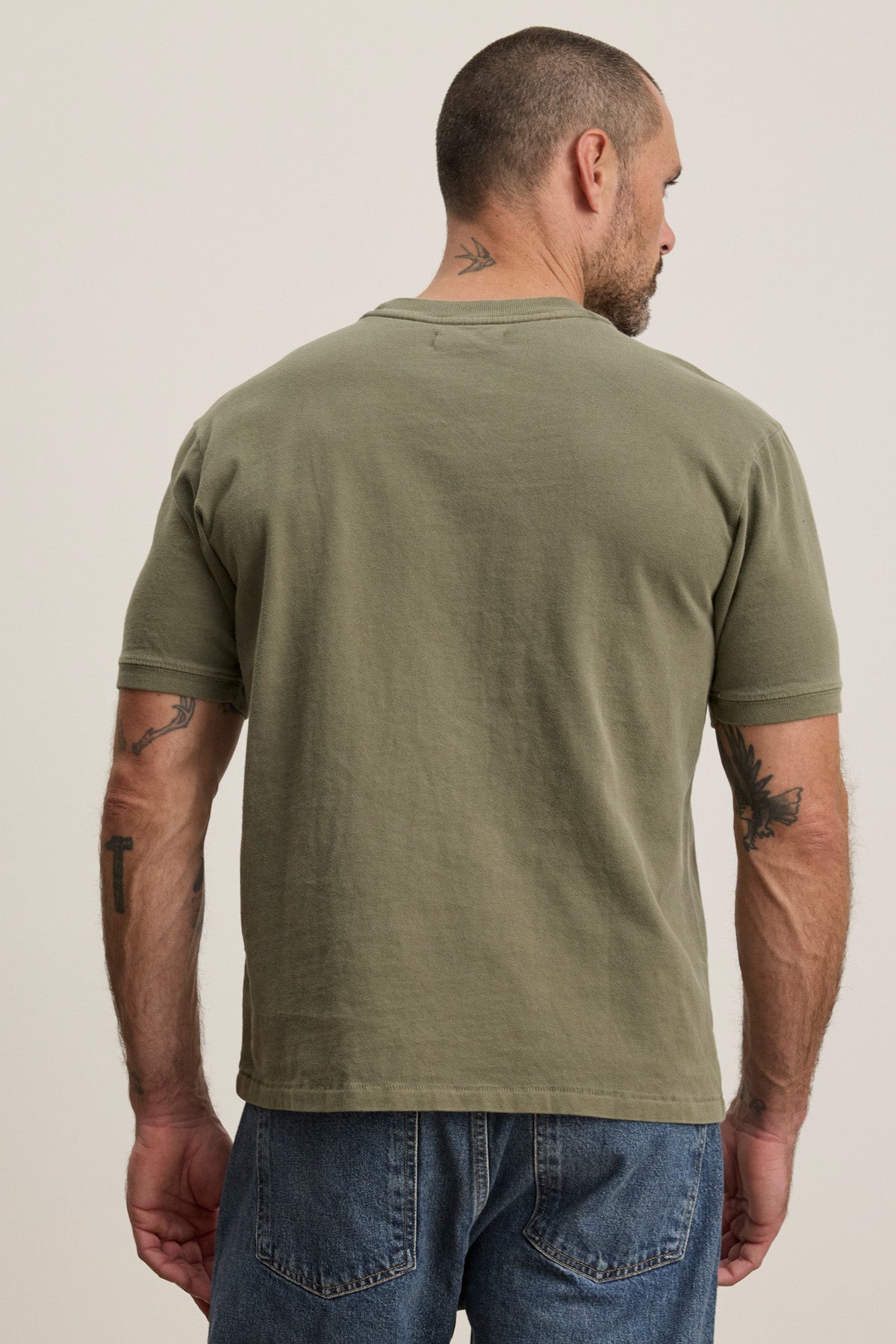 A man with short hair and tattoos stands with his back to the camera, wearing the ELIAS TEE by Velvet by Graham & Spencer in olive green and blue jeans.